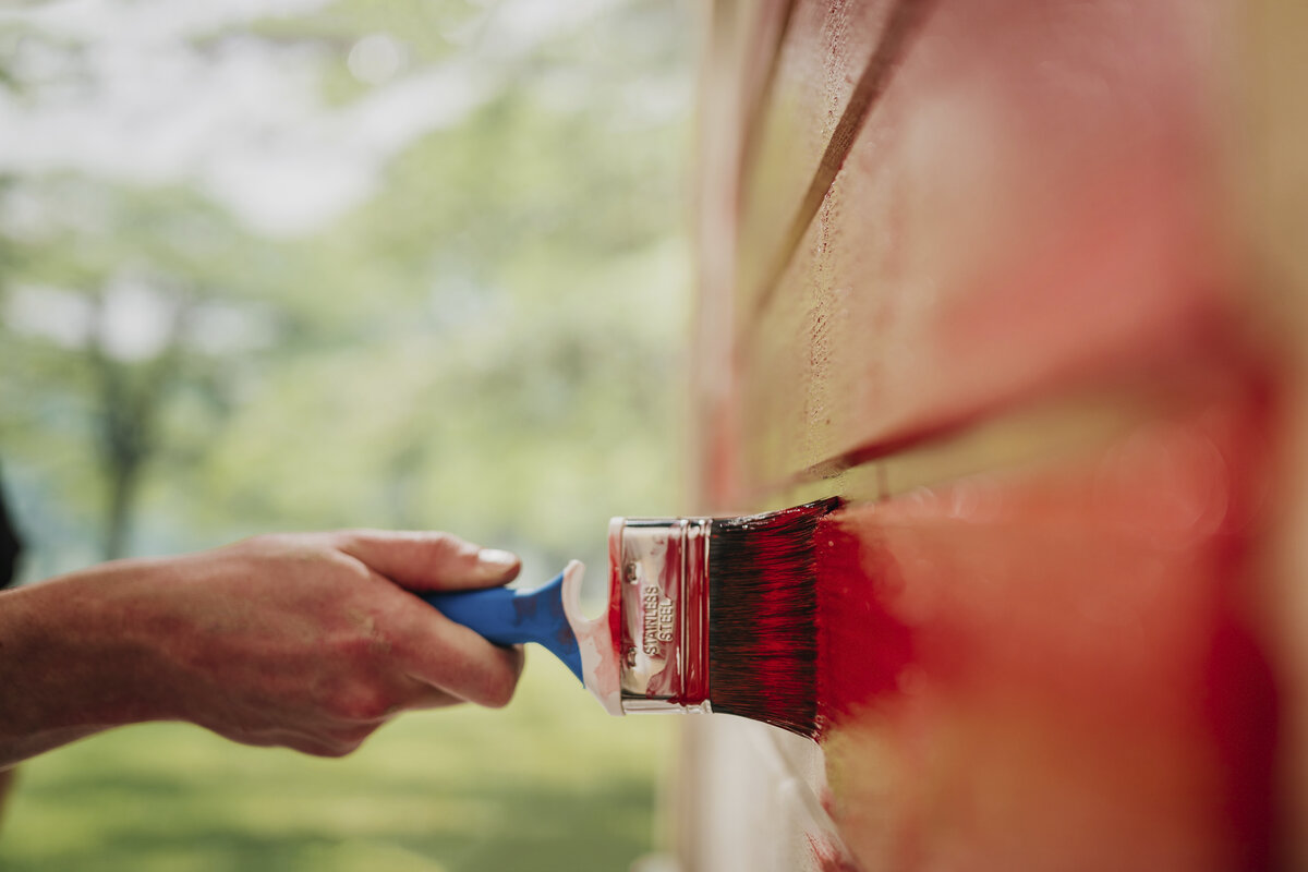 Kuvassa näky pensselillä ulkoseinää punaiseksi maalaava käsi.